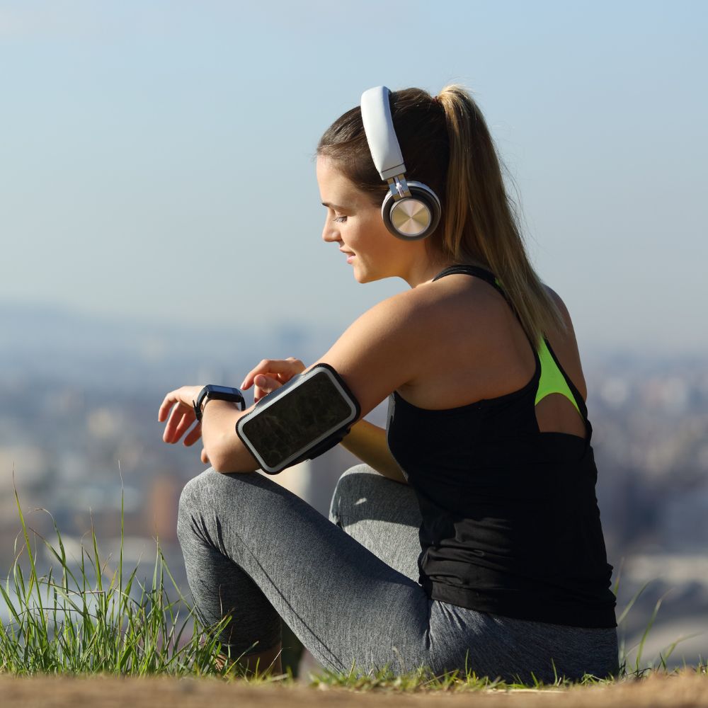 Woman sitting outdoors with headphones and fitness tracker, overlooking a cityscape.