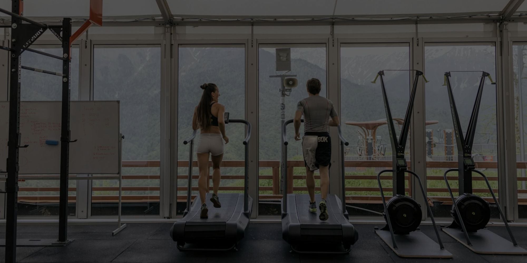 Two people using exercise bikes in a gym with large windows showing an outdoor view.